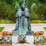 Pomnik - klęczący święty Jan Paweł II. Przed pomnikiem dwa klęczniki. Na drugim planie ławki, drzewa i trawnik.