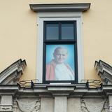 Od dołu ozdobny gzyms, nad którym w prostokątnym oknie murowanego budynku, widnieje duże zdjęcie do połowy papieża Jana Pawła II. W czerwonej pelerynie, pod spodem w białej albie, z białą piuską na głowie.