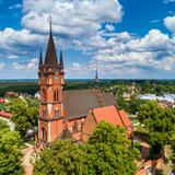 Widok na cały kościół z wysoka wieżą i boczna kaplicą, dawnym kościołem długoszowskim. Dookoła zabudowa wsi i dużo zieleni.