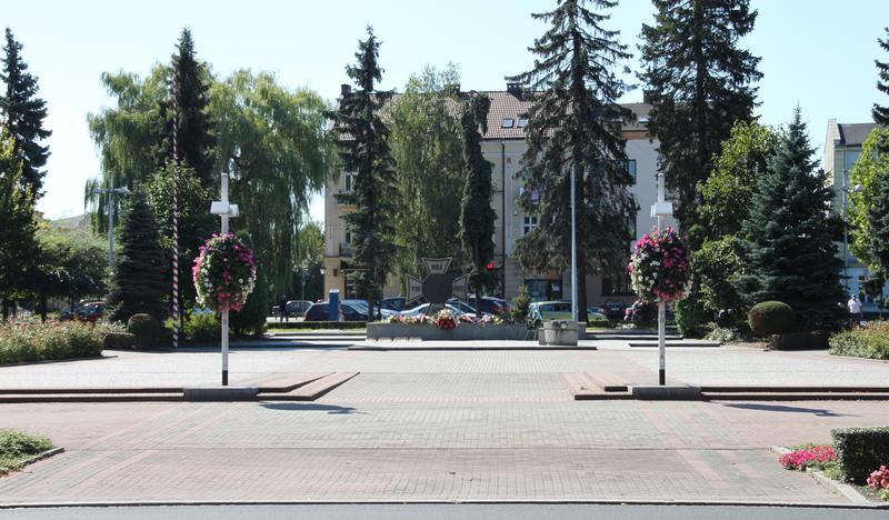 Na zdjęciu znajduje się Plac Tadeusza Kościuszki w Oświęcimiu, gdzie odbywają się różne uroczystości. W oddali widać Grób Nieznanego Żołnierza, wokół kwiaty, z tyłu budynek za drzewami.