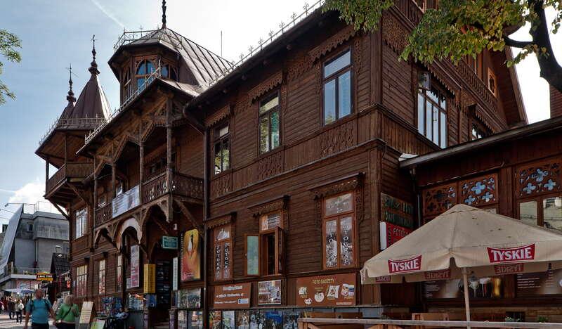 Duża drewniana willa w stylu alpejskim, piętrowa, ze zdobionym balkonem na piętrze, nad wejściem. Przed willą kilka osób, po prawej stronie parasol z napisem Tyskie.