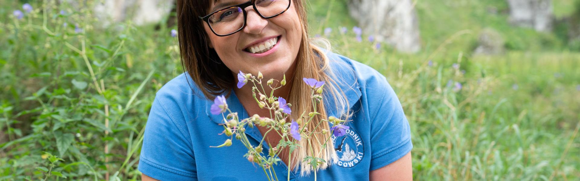 Uśmiechnięta kobieta w błękitnej koszulce z polnym kwiatem w ręce na tle skałek jurajskich