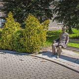 Brukowana ulica. Dalej wśród zieleni stoi ławeczka z posągiem siedzącego Karola Wojtyły. Po lewej tuje, za nimi fragmenty budynku częściowo zasłoniętego przez rosnące, wysokie drzewa iglaste.