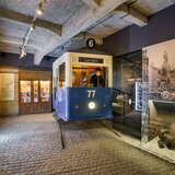 Wagonik tramwajowy w sali muzealnej w Fabryce Emalia Oskara Schindlera. Sufit jest betonowy, posadzka brukowana, a na ścianach wiszą zdjęcia.