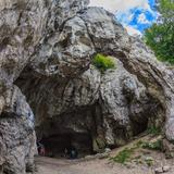Wejście do jaskini - duży otwór w skale. Obok drzewa wysokie i fragment nieba z białymi chmurami. W jaskini stoi trzech turystów.