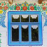 Okno drewnianego domu w niebieskich ramach, ozdobione wycinankami w kształcie okrągłych serwetek, dom malowany w zalipiańskie wzory.