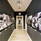 Czarny korytarz w muzeum. Na ścianach wiszą czarno-białe zdjęcia, przedstawiające różnych ludzi.