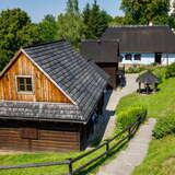 Drewniane chaty w Muzeum PTTK w Dobczycach. Do chat prowadzą schodki, a wokół nich znajduje się sporo zieleni.