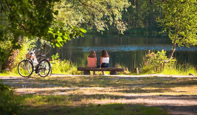 Dwie dziewczyny siedzą na ławce nad stawem w lesie. Obok stoją zaparkowane rowery.